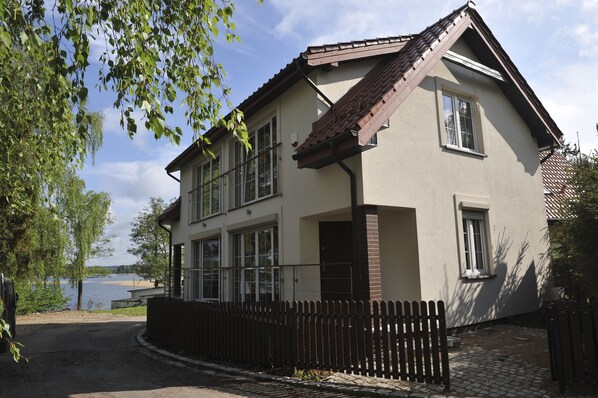 A two-story house with a brown roof, beige walls, and a wooden fence. - Vertigo Narie (Morag)