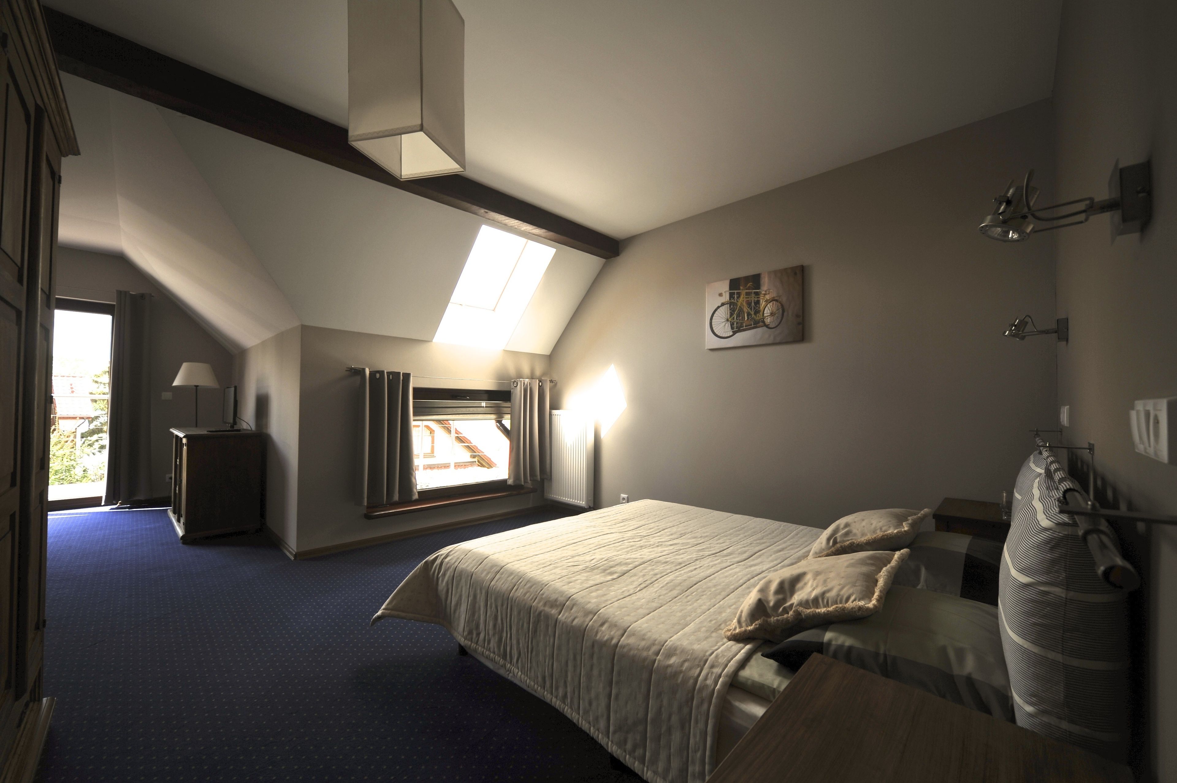 A bedroom with a large bed, a desk, a chair, a radiator, a window with curtains, and a bicycle artwork on the wall.