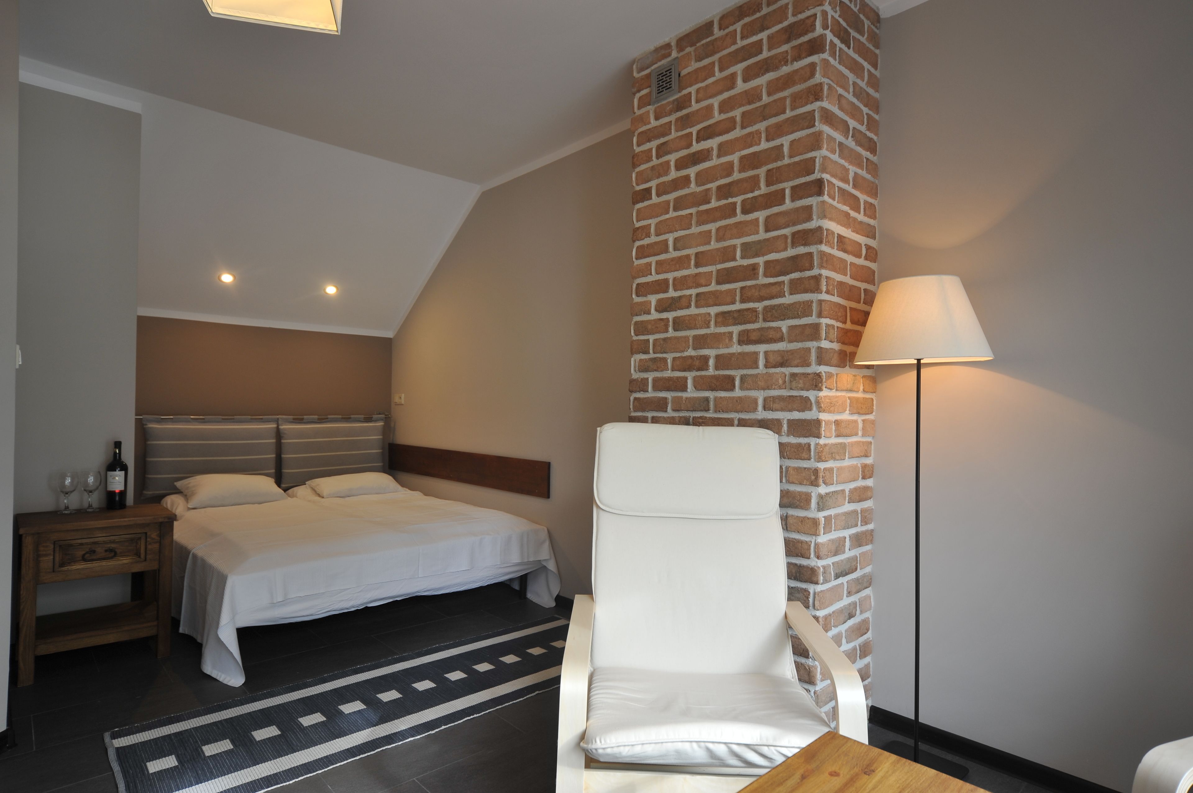A bedroom with a bed, a nightstand, a chair, a lamp, and a brick wall.