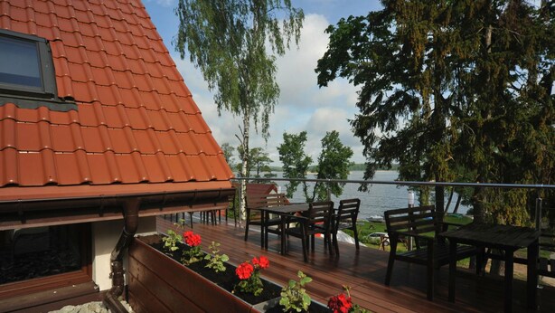 A wooden deck with outdoor seating and flower planters, overlooking a body of water and trees. - Vertigo Narie (Morag)