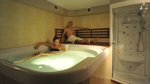 A bathtub with two people sitting inside, a shower area to the right, and a wooden bench against the wall. - Vertigo Narie (Morag)