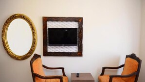 Una habitación con dos sillas de madera, una mesita, un televisor montado en la pared y un espejo grande.