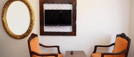 Una habitación con dos sillas de madera, una mesita, un televisor montado en la pared y un espejo grande.