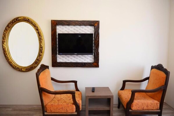 Una habitación con dos sillas de madera, una mesita, un televisor montado en la pared y un espejo grande.