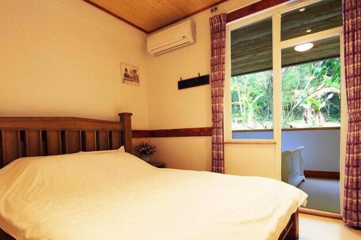 A bedroom with a wooden bed, a nightstand, a window with curtains, and an air conditioning unit.