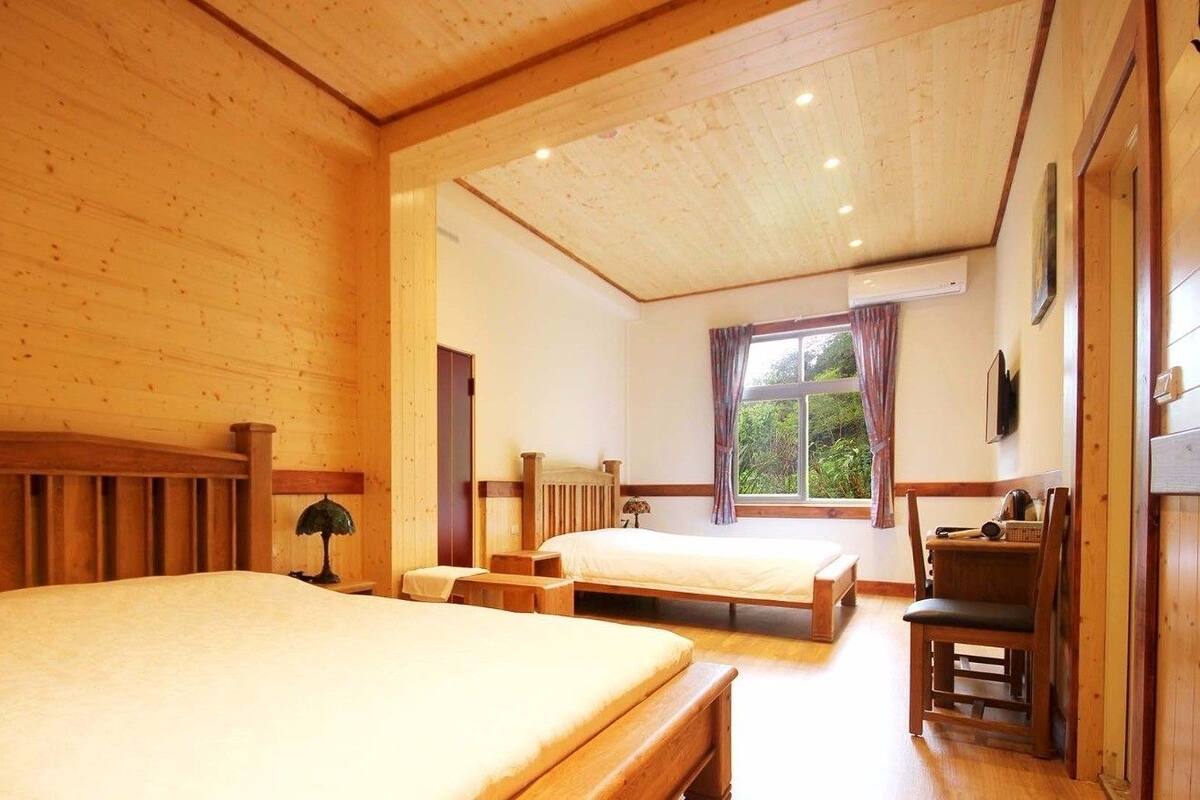 A wooden-paneled room with two single beds, a small table, and a window with curtains.