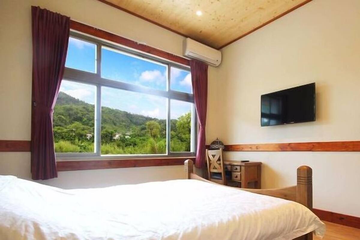 A bedroom with a large bed, a TV mounted on the wall, and a window offering a view of a green landscape.