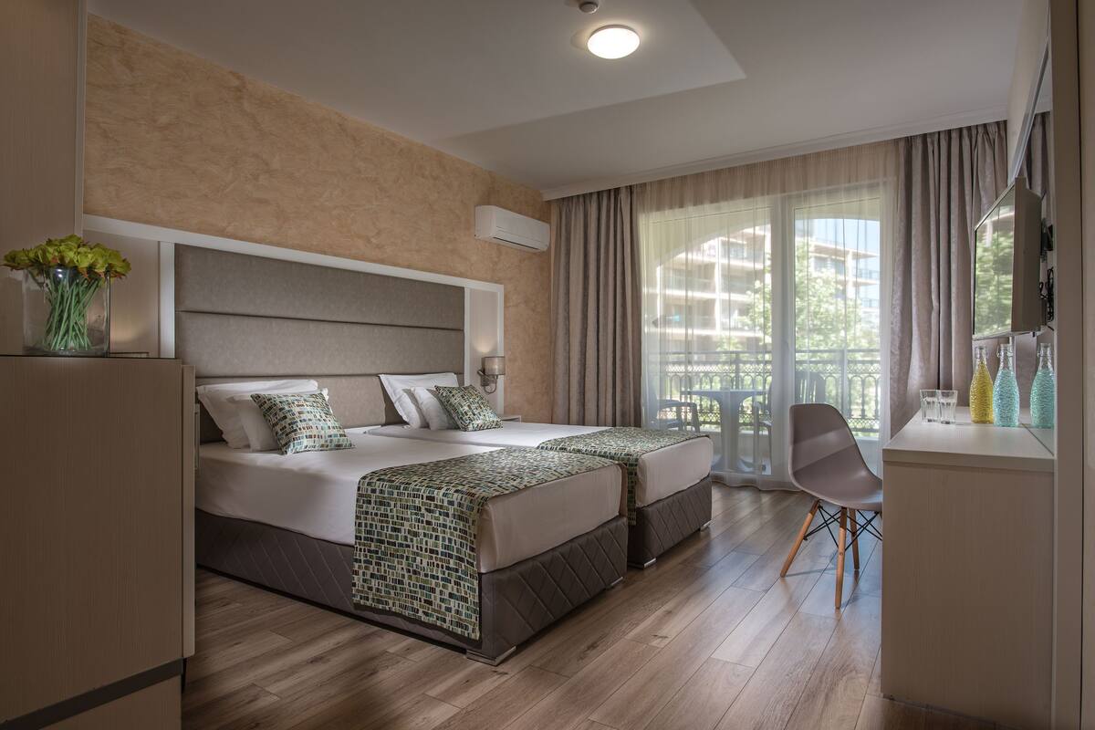 Une chambre d’hôtel avec un grand lit, deux tables de chevet, une chaise, un bureau et une fenêtre avec des rideaux.