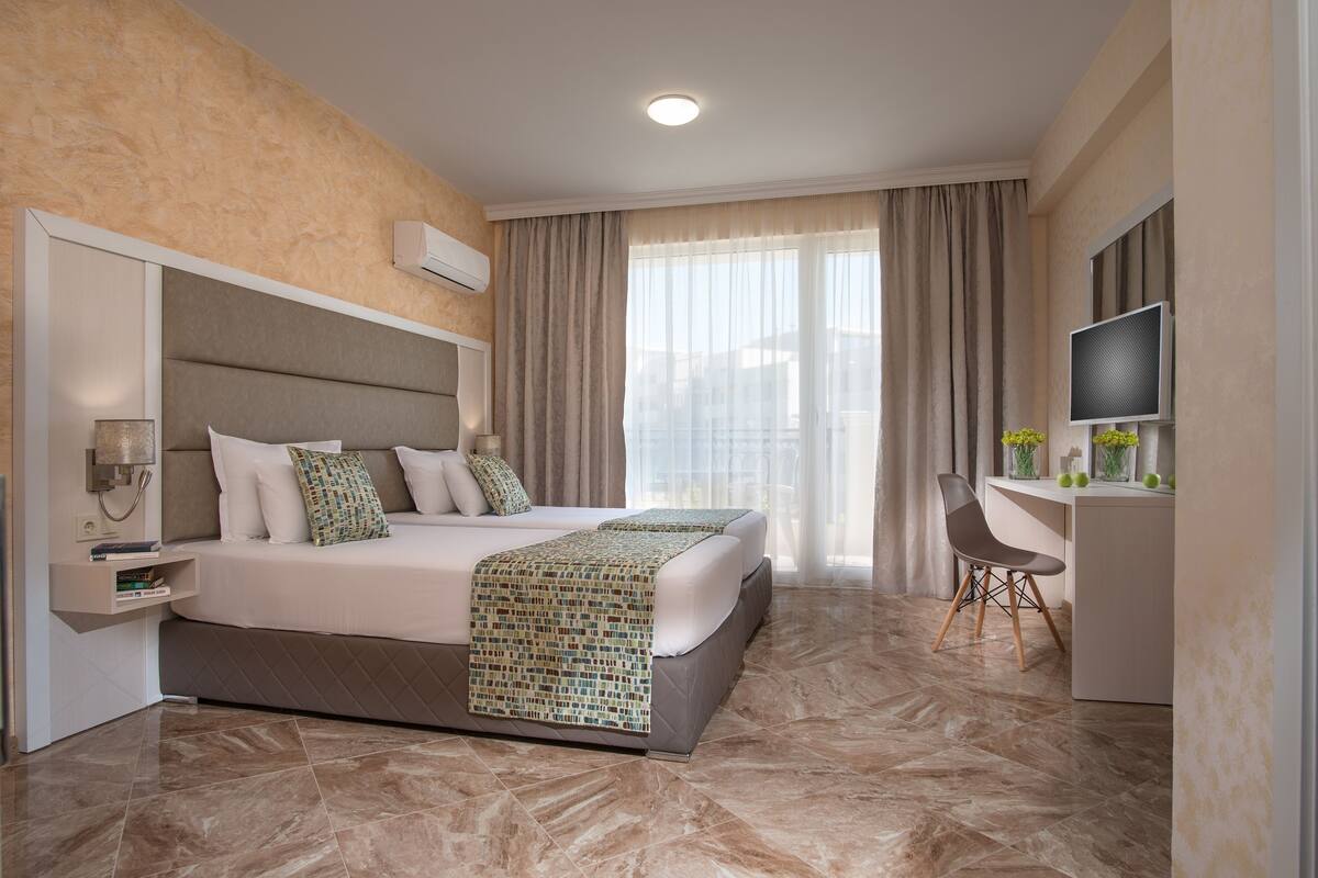 Une chambre d’hôtel avec un grand lit, un bureau avec une chaise, un téléviseur à écran plat et une grande fenêtre avec des rideaux.