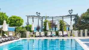 Un espace piscine avec des chaises longues et des parasols, une pergola agrémentée de lampes et une vue sur la mer.