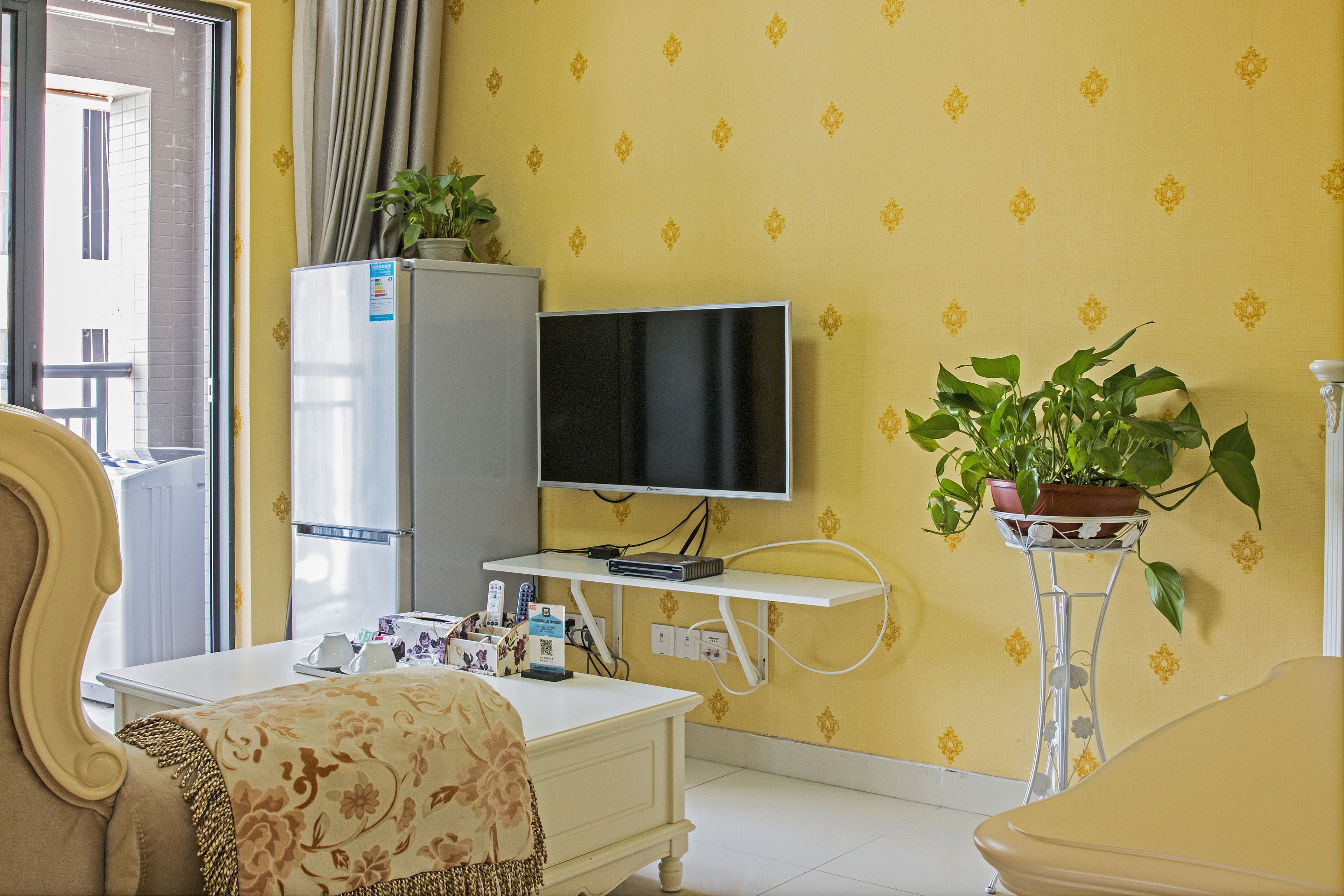 Een kamer met een televisie, een koelkast, een wit bureau, een kamerplant en een gestreept bankje.