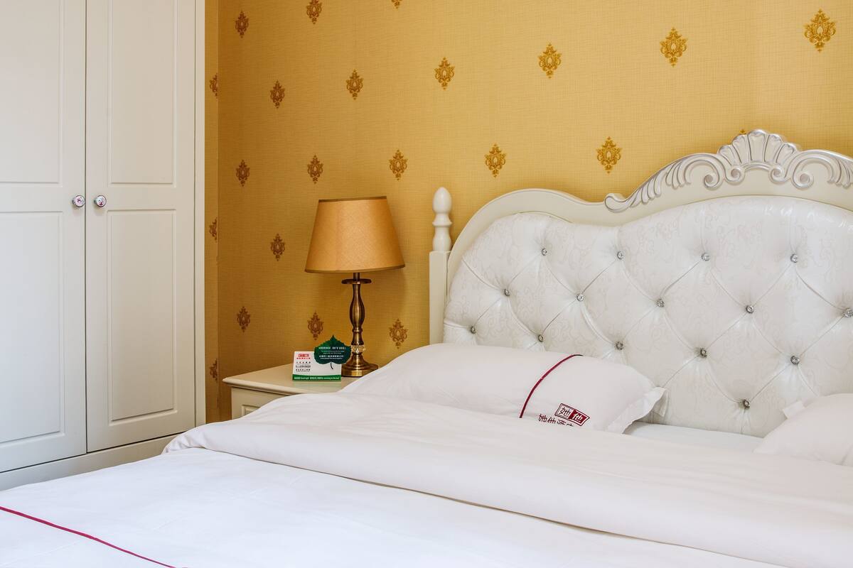 Un letto ben rifatto con lenzuola bianche e un tocco di colore rosso, una testiera bianca con motivo capitonné, un comodino con una lampada e una scatola, e una parete con carta da parati decorata.