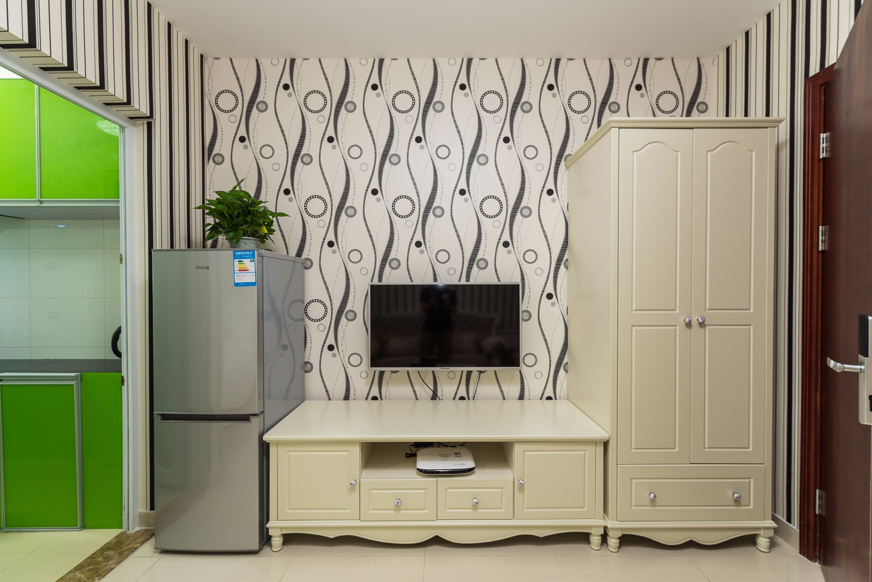 A compact kitchen with a refrigerator, a TV mounted on the wall, and a cabinet with drawers.