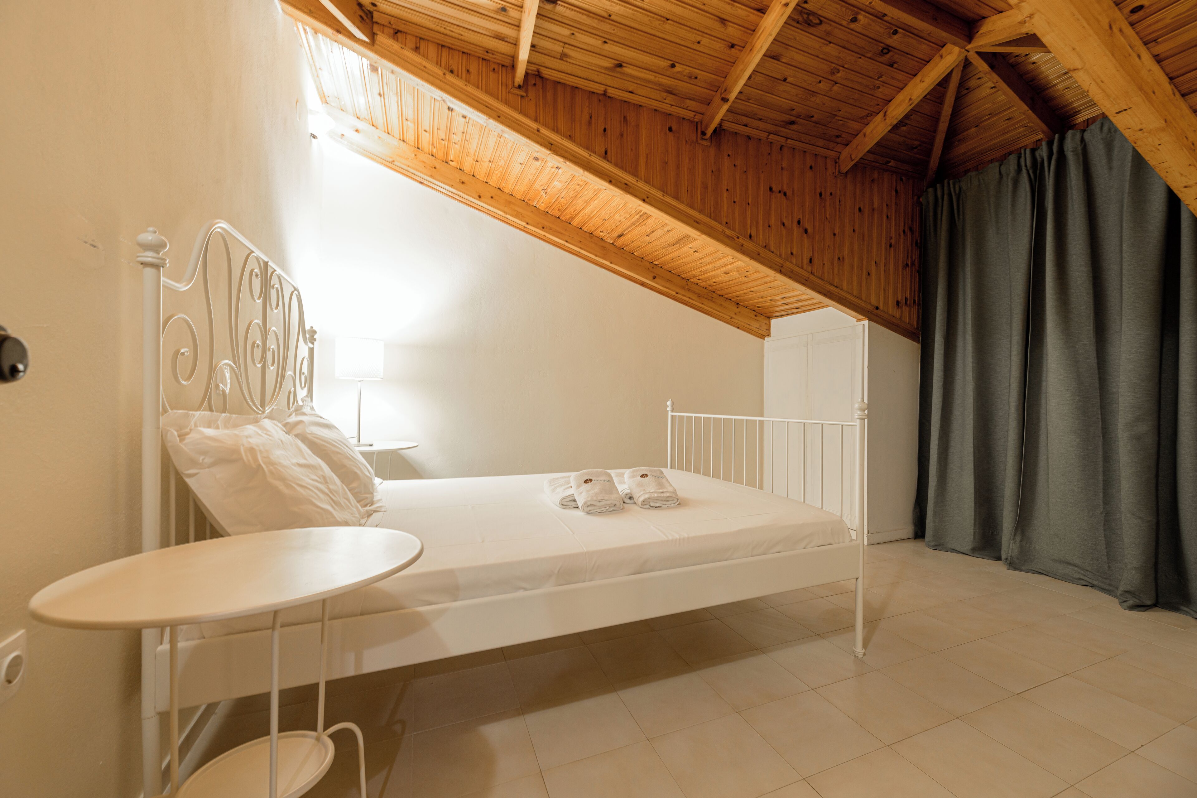 Camera singola con un letto, soffitto in legno, un tavolino rotondo e una finestra con tende.