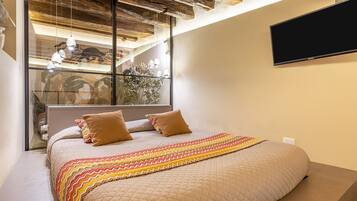 Ein Schlafzimmer mit einem großen Bett, einer Holzbalkendecke, einem an der Wand montierten Fernseher und einem Fenster mit Blick auf eine Treppe und Pflanzen.