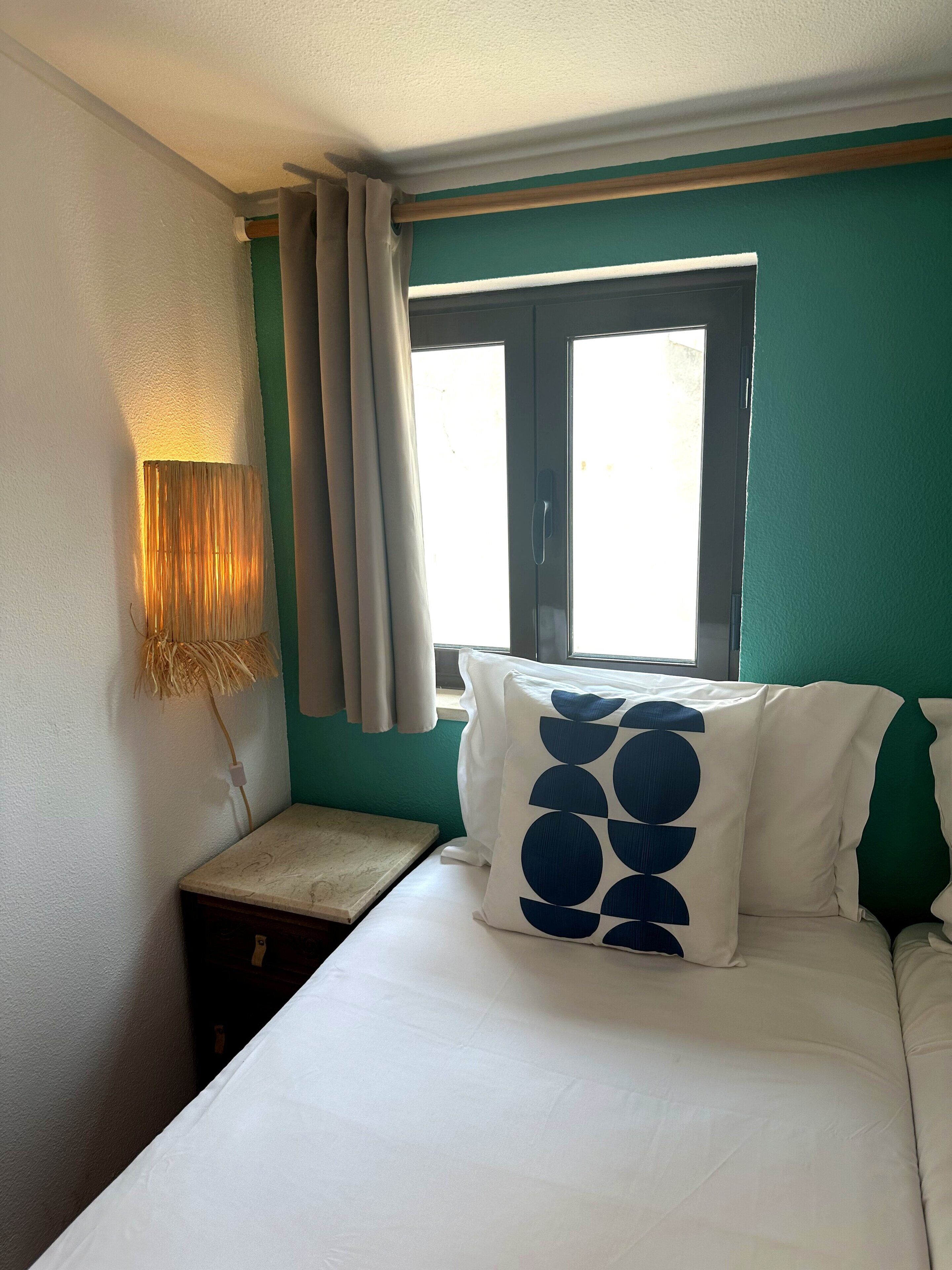 Une chambre d’hôtel avec un lit, une table de chevet, une lampe, une fenêtre avec des rideaux et un mur vert.