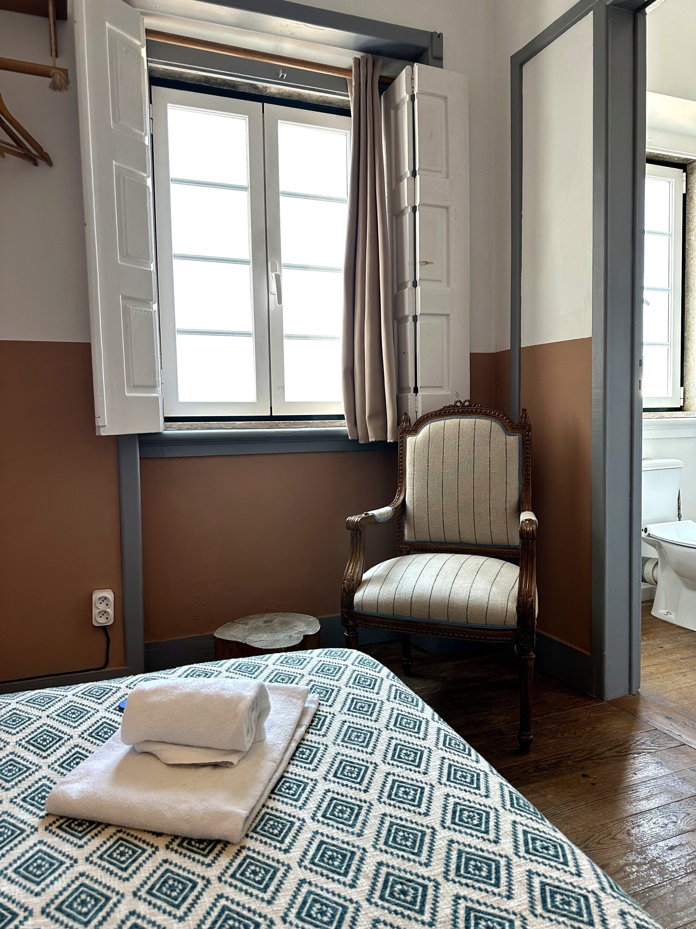 A room with a bed, a chair, a window with curtains, a wooden floor, and a wall socket.