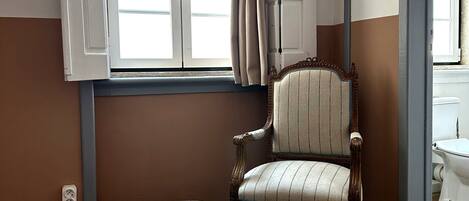 Una habitación con cama, una silla, ventana con cortinas, piso de madera y un enchufe.