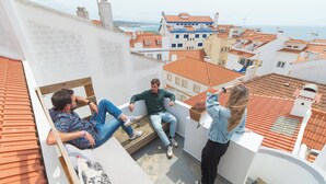 A rooftop terrace with three people sitting and one standing, overlooking a coastal town with terracotta roofs.