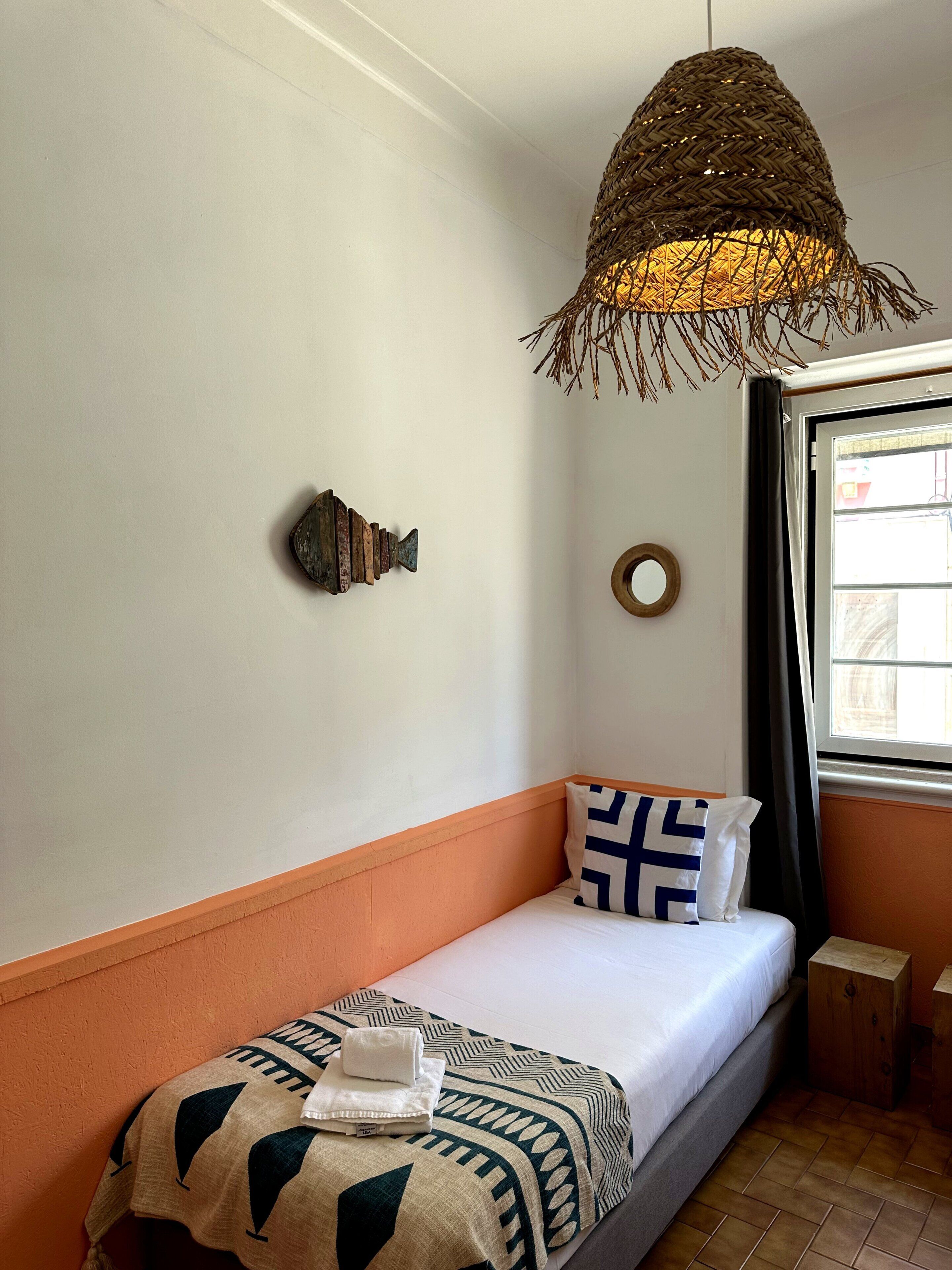 A single bed with a patterned blanket, a hanging woven light fixture, a wall-mounted decorative object, and a window with curtains.