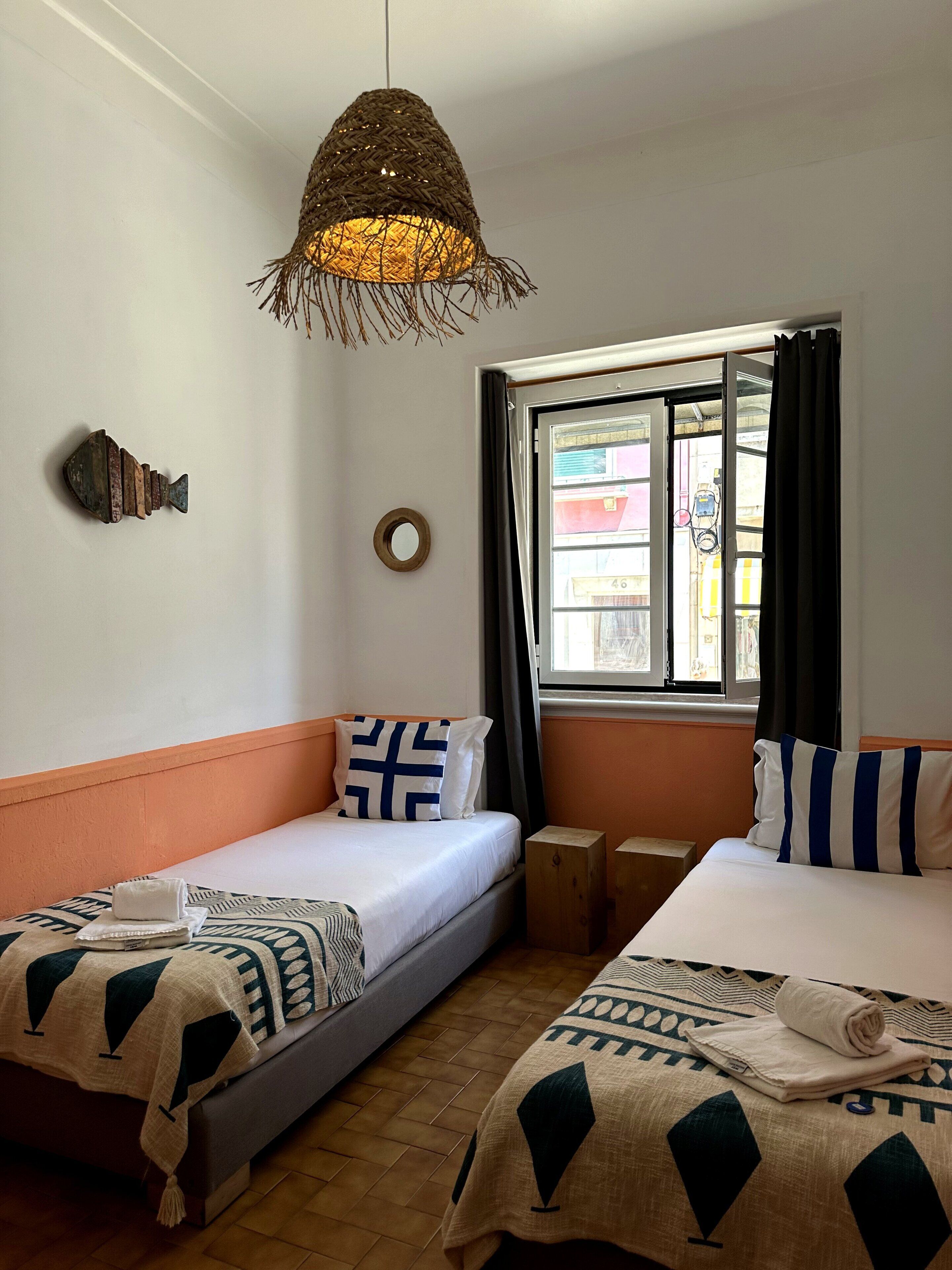 A hotel room with two beds, a hanging light fixture, a fish decoration, a window with curtains, and a view of the outside.