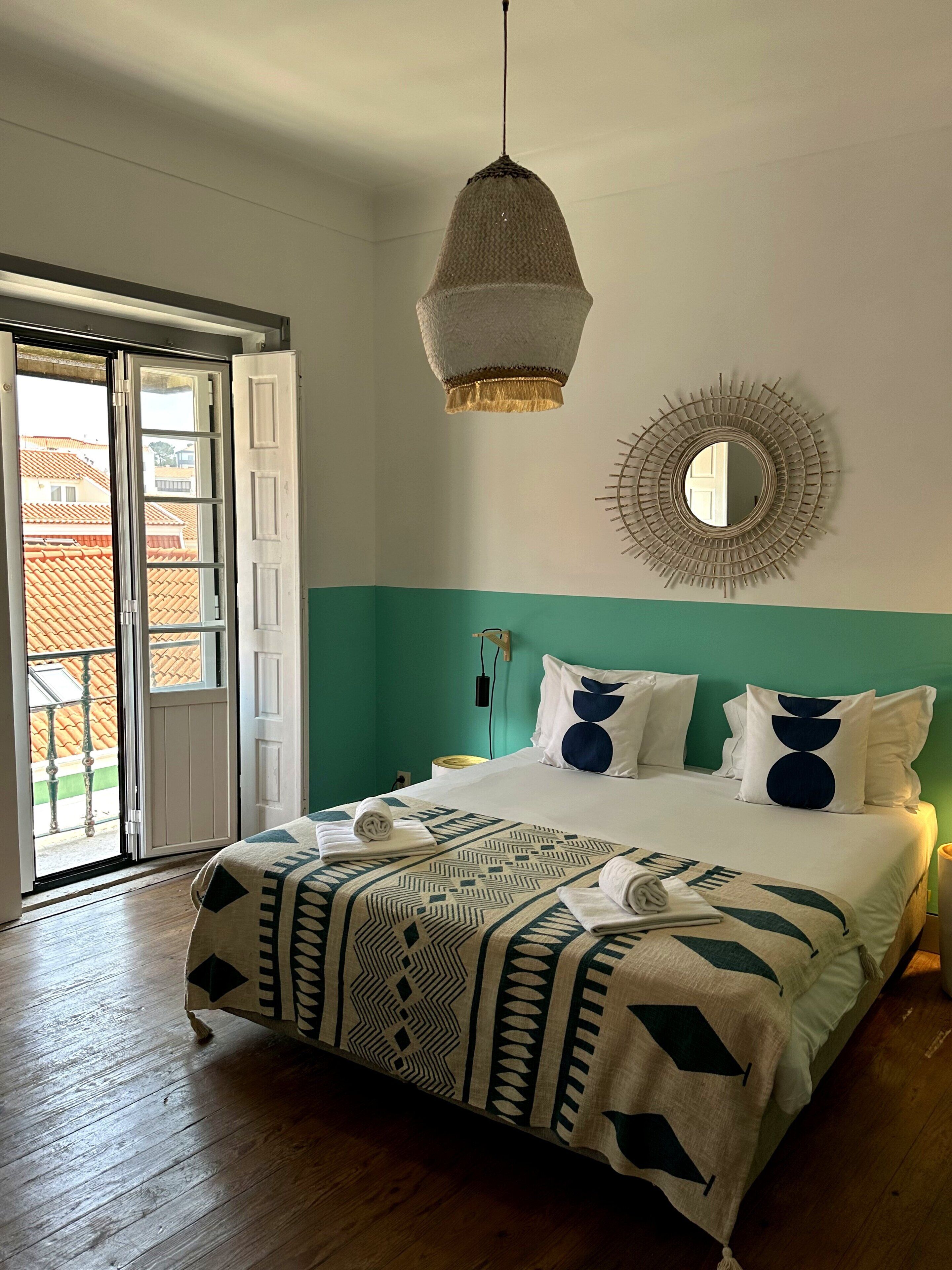Une chambre d’hôtel avec un lit, un luminaire suspendu, un miroir mural et une vue sur l’extérieur, visible à travers une porte.