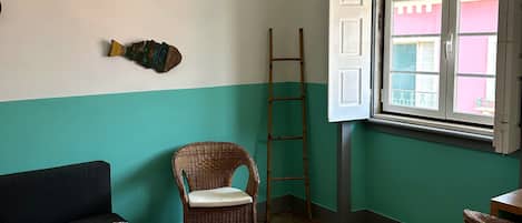 A room with a wooden floor, a wicker chair, a sofa, a hanging light fixture, and a window with open shutters.
