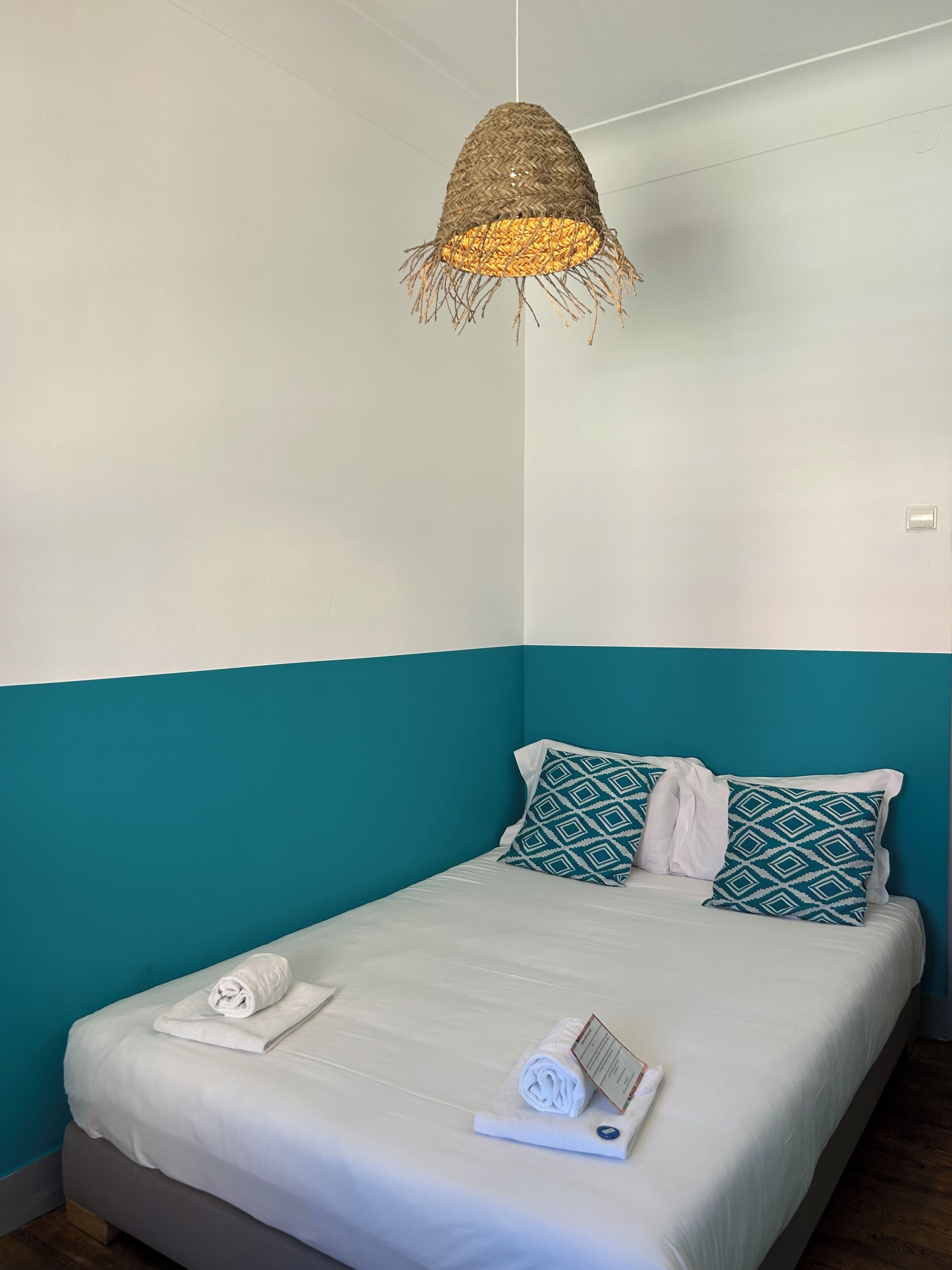 A single bed with white bedding, teal accent walls, and a woven pendant light fixture.