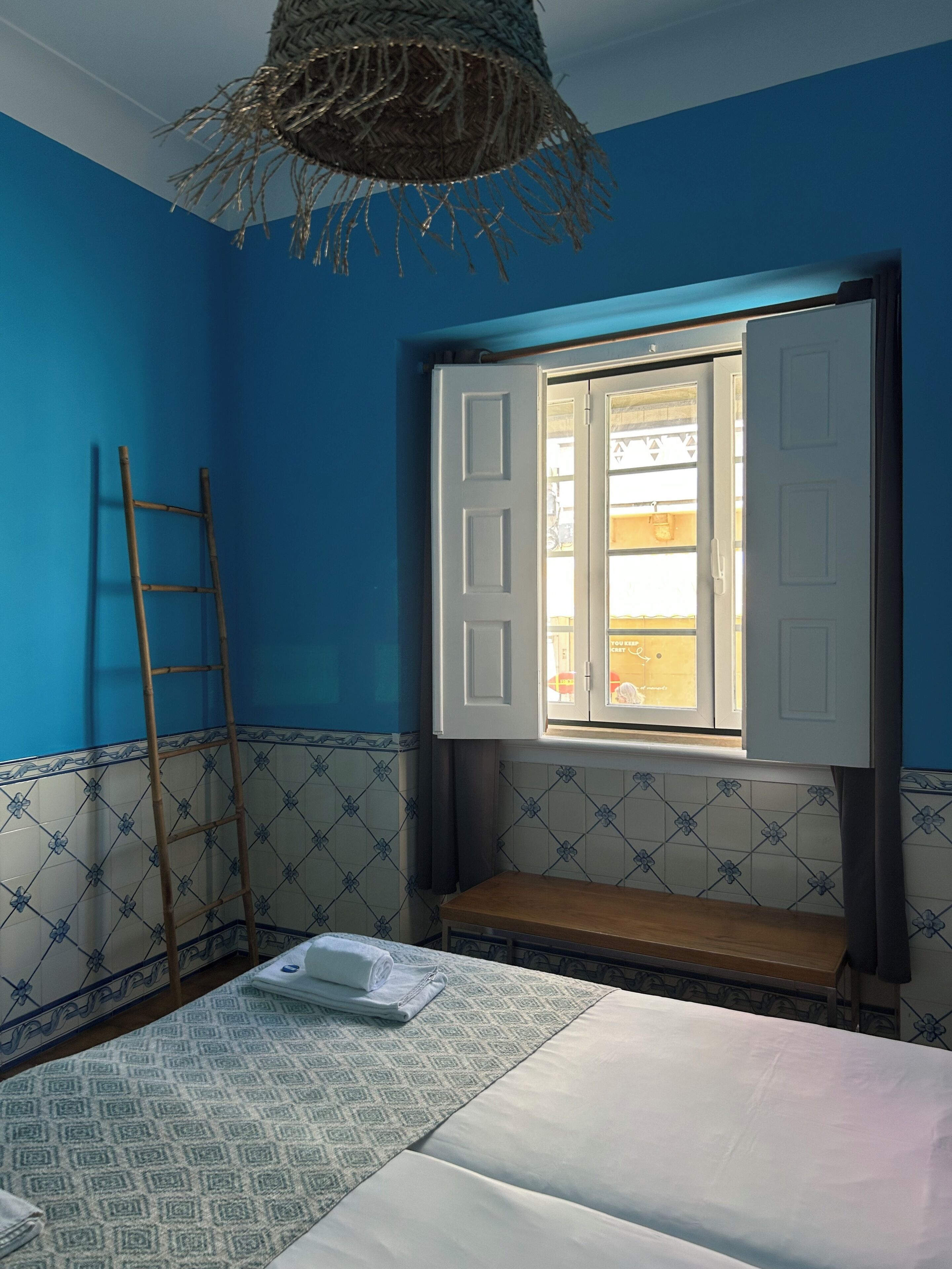 A room with a bed, a window with open shutters, a ladder, and a patterned wall.