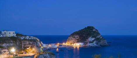 A coastal town at dusk with buildings along the cliffside and a prominent rock formation in the sea.