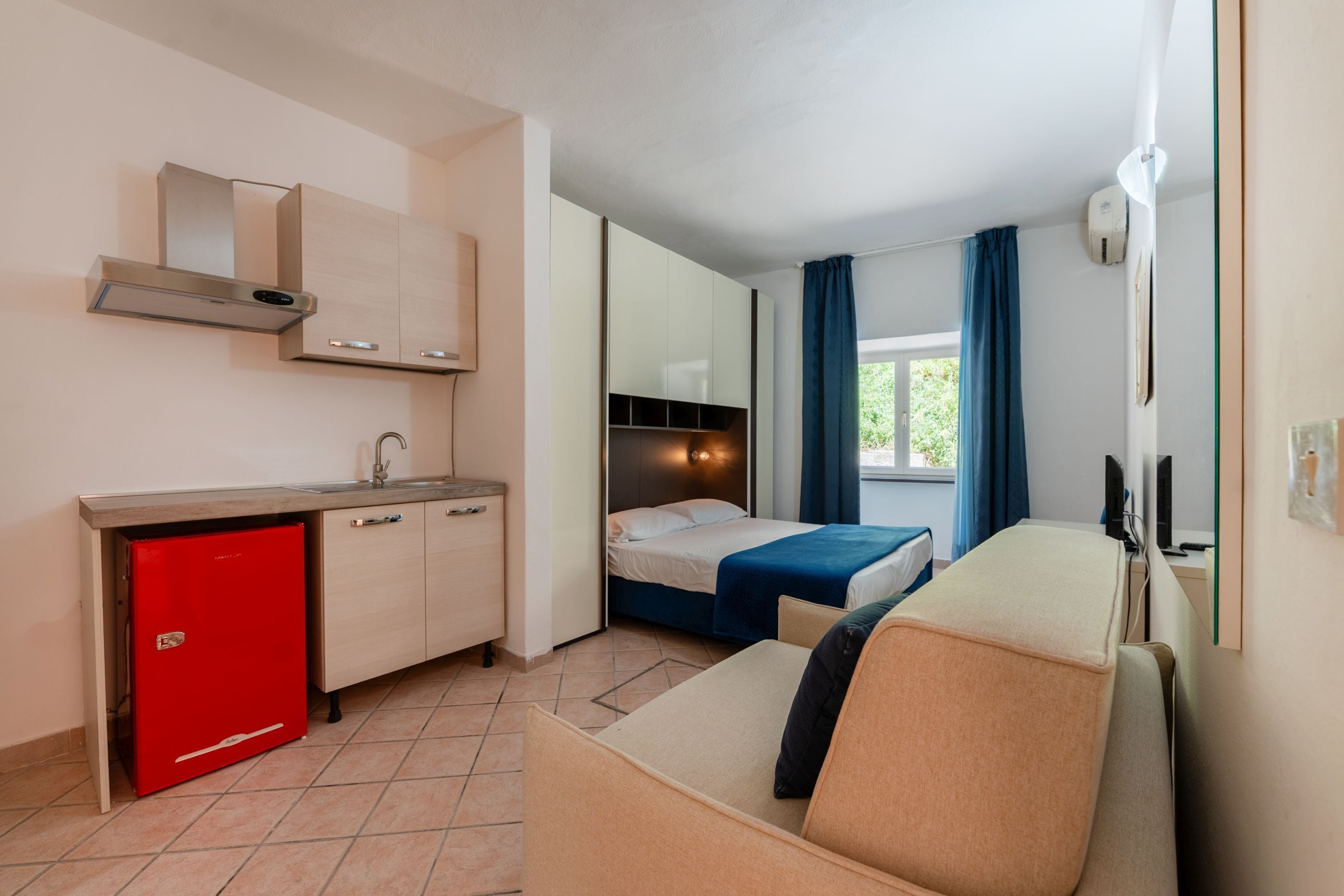 Une chambre d’hôtel compacte comprenant un coin cuisine, un lit, un canapé et une fenêtre avec des rideaux.