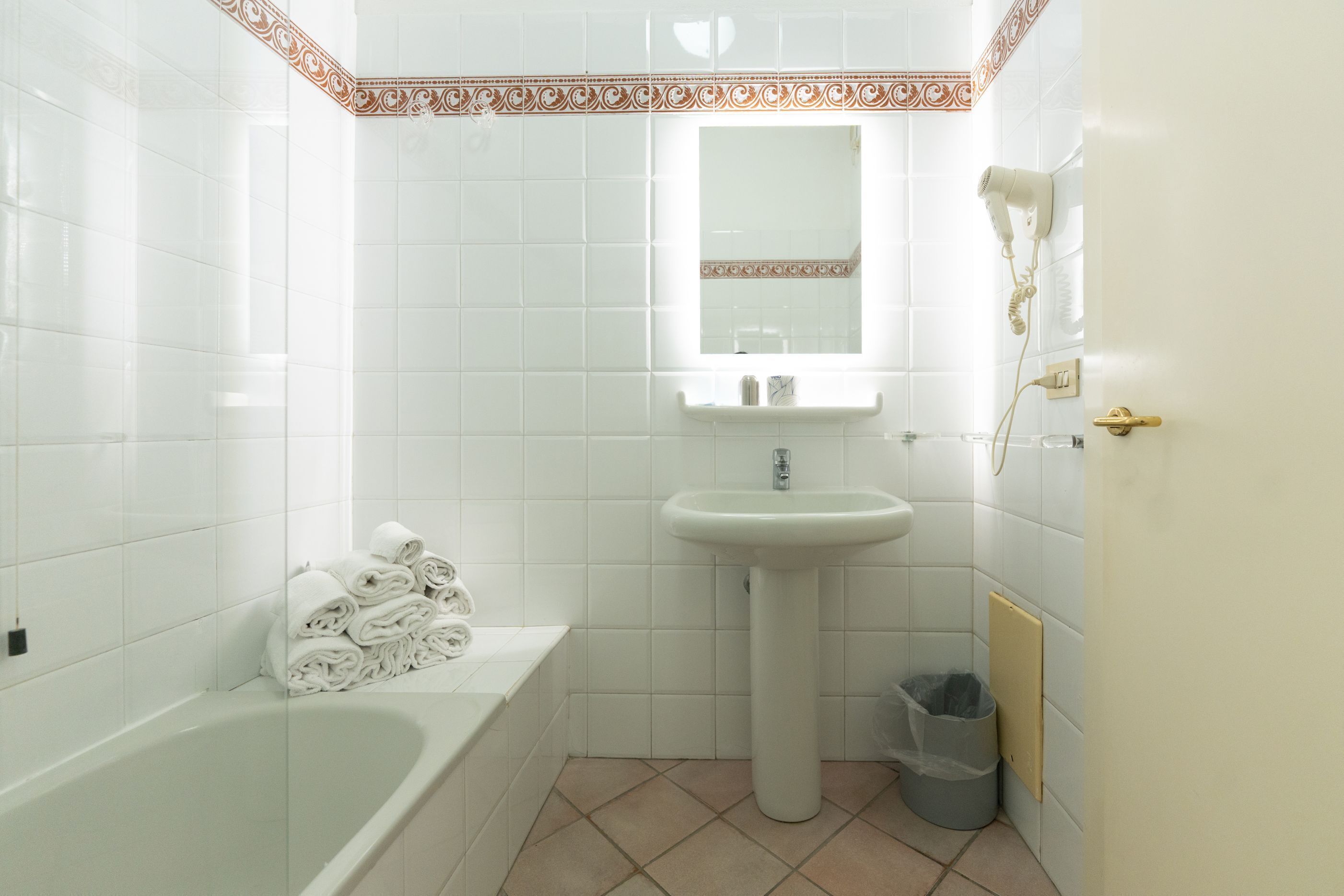 A bathroom with white tiles, a sink, a mirror, a hairdryer, a trash bin, and a stack of towels.