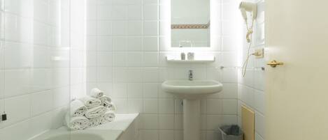 A bathroom with white tiles, a sink, a mirror, a hairdryer, a trash bin, and a stack of towels.