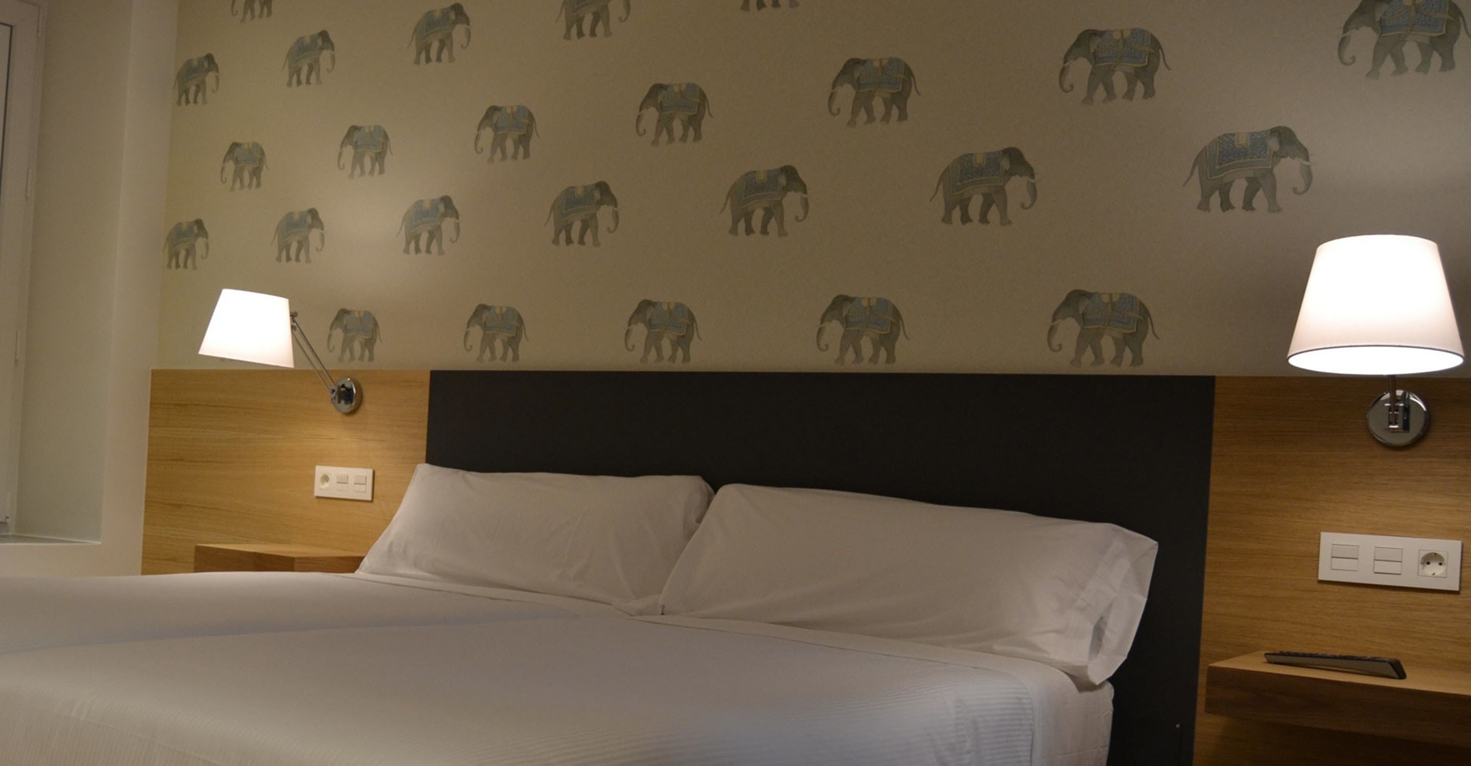 A hotel room with a bed, two bedside lamps, a nightstand, and a wall with elephant patterns.