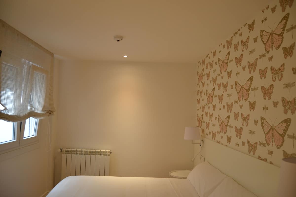 A bedroom with a bed, a window with curtains, a radiator, a wall-mounted lamp, and a wall with butterfly decals.