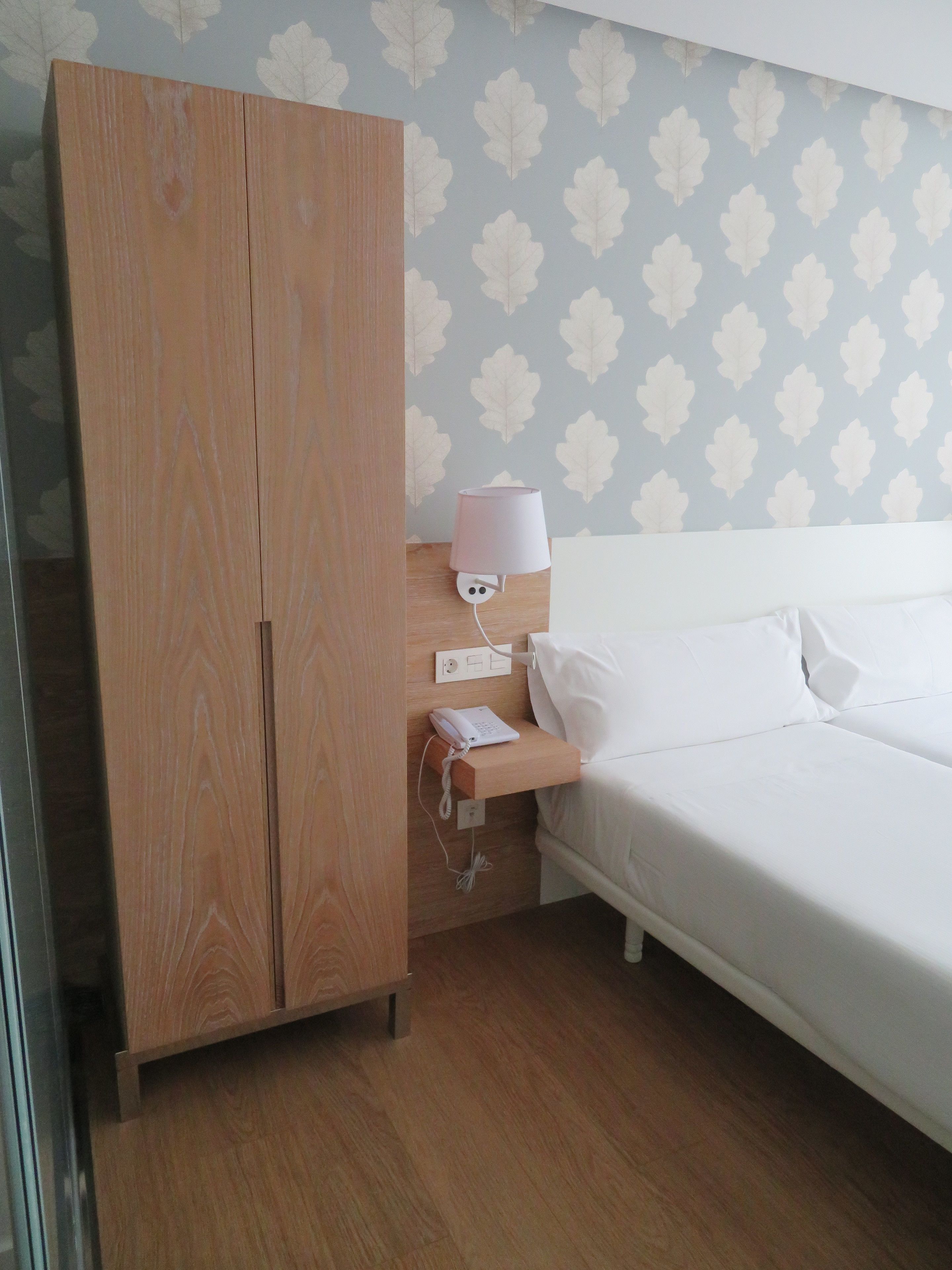 A hotel room with a wooden wardrobe, a bedside table with a lamp, a phone, and a neatly made bed.