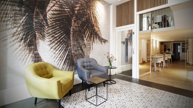 Un vestíbulo moderno con un gran mural de hoja de palma, un sillón amarillo, un sillón gris y una pequeña mesa con flores.