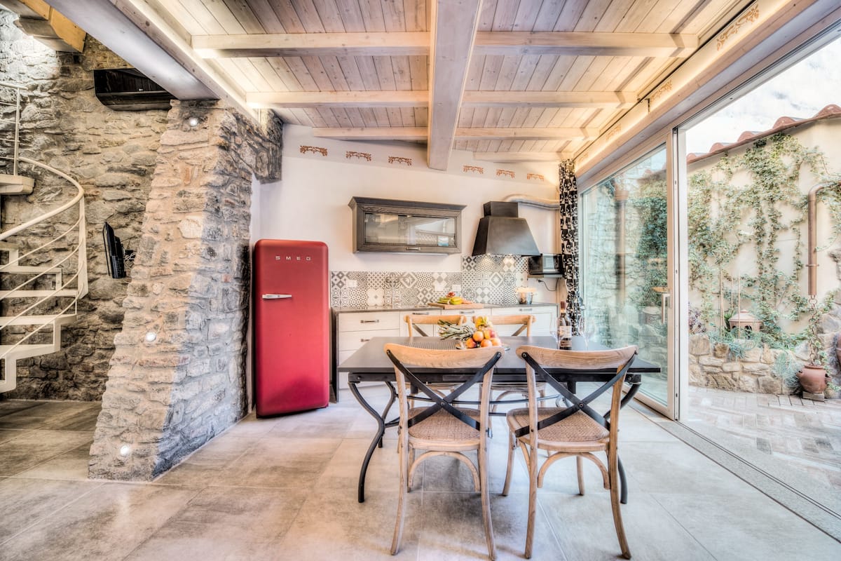 A kitchen with a stone wall, a red refrigerator, a wooden dining table with chairs, and a large window with a view of greenery outside.