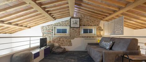 Un dormitorio acogedor en el ático con vigas de madera a la vista, paredes de piedra, un sofá, un televisor y una ventana con persianas.