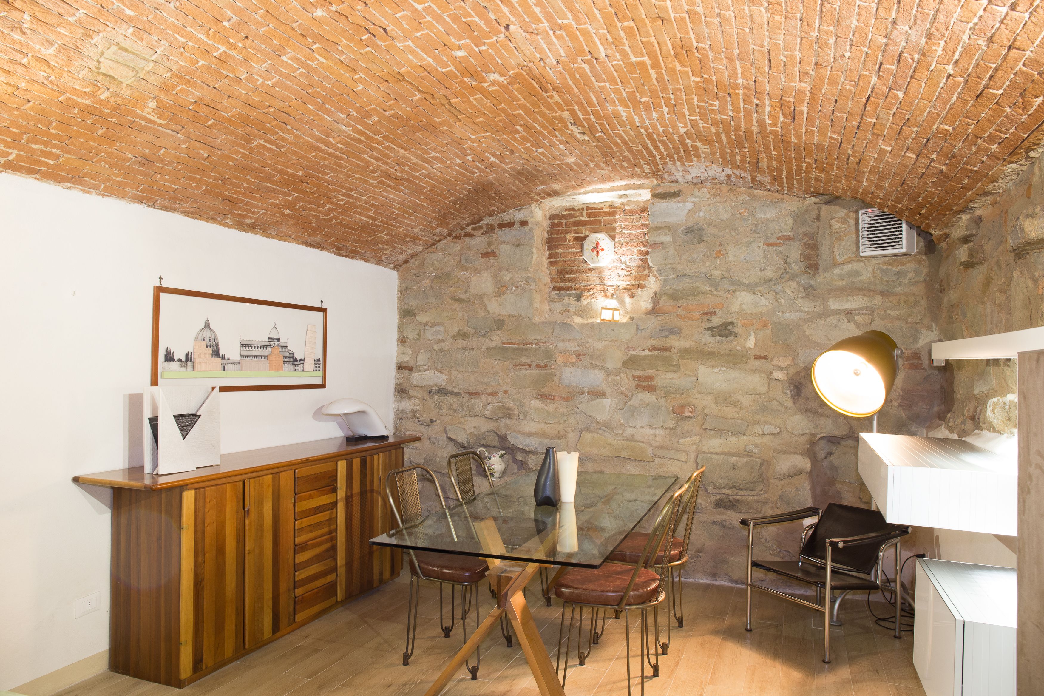 Un comedor con una mesa de vidrio, sillas de metal, un aparador de madera y una pared de piedra.