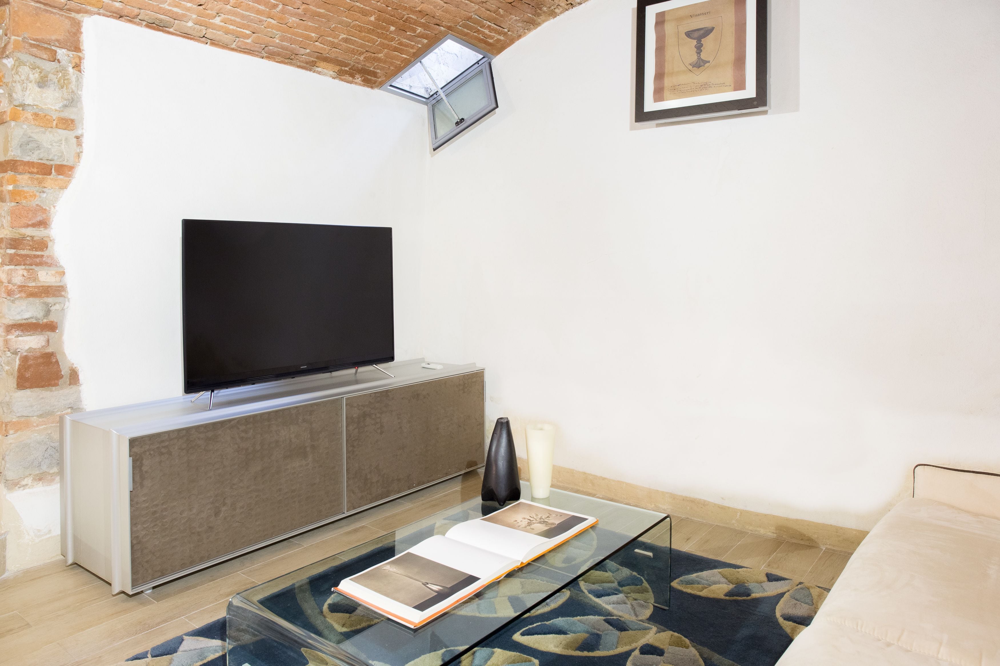 A modern living room with a flat-screen TV on a cabinet, a glass coffee table with a book, a candle, and a framed picture on the wall.