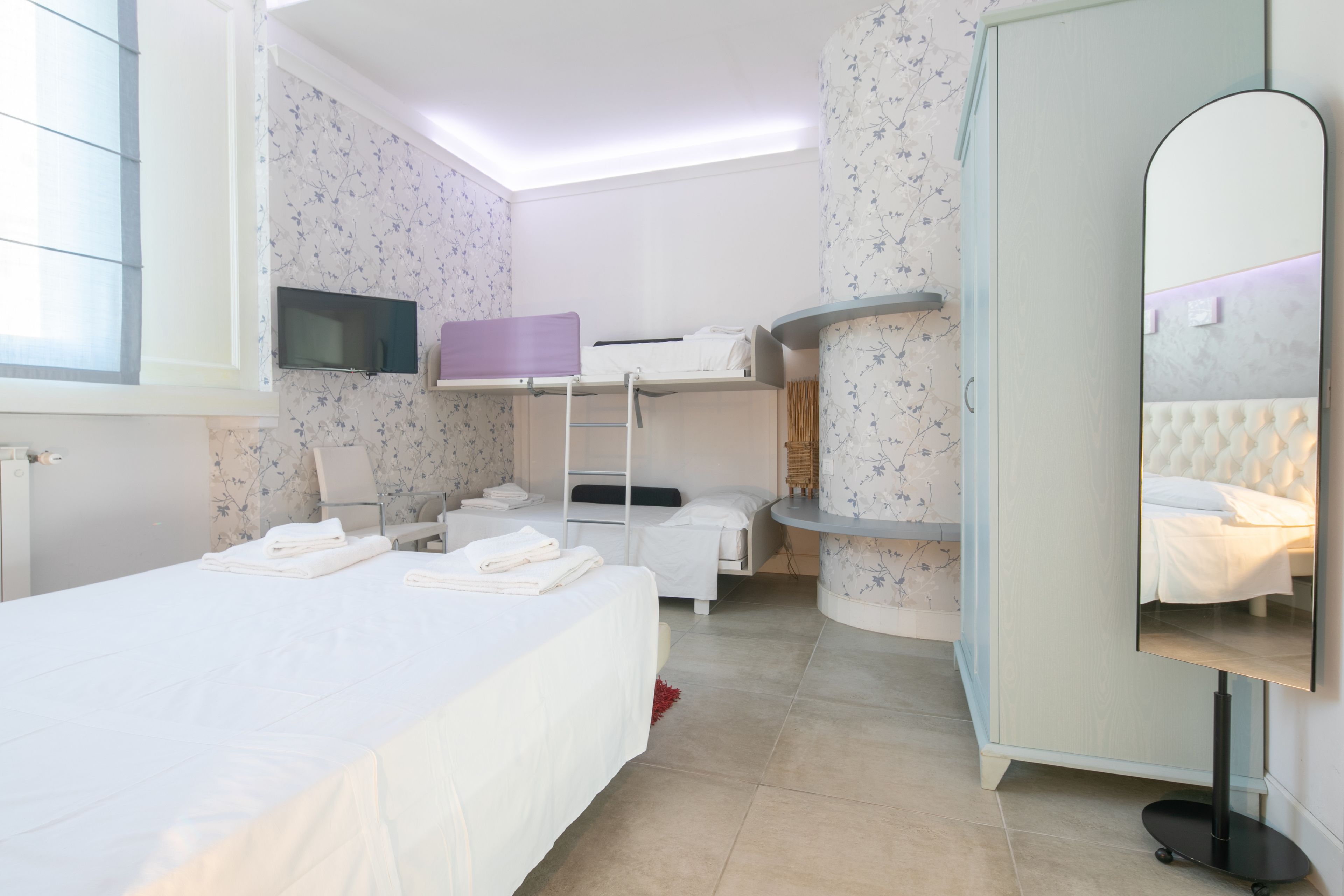 Une chambre d’hôtel avec un lit superposé, un téléviseur à écran plat fixé au mur, un grand miroir et une table de chevet avec une lampe.