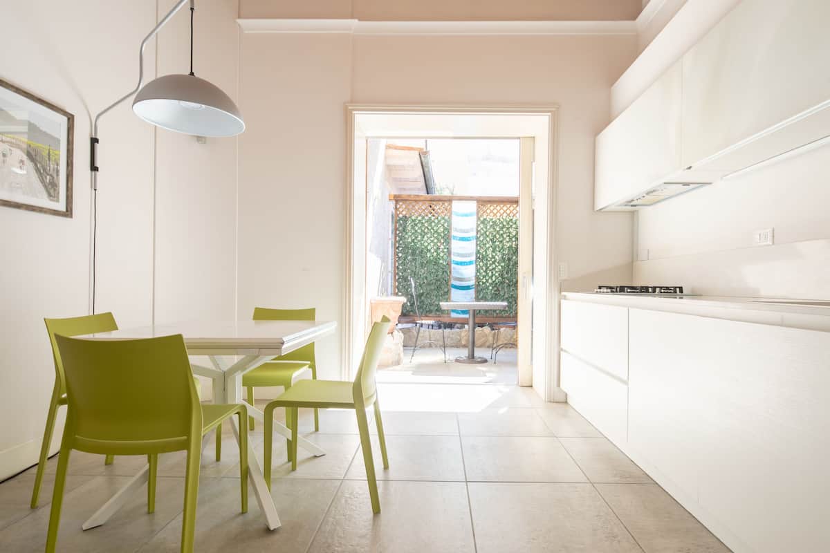 A modern kitchen with white cabinets, a dining table with green chairs, and a view of a garden through an open door.