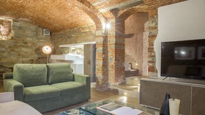 A living room with a green sofa, a glass coffee table with a book, a TV, and stone walls. - Giglio Apartments by Mmega (Florence)