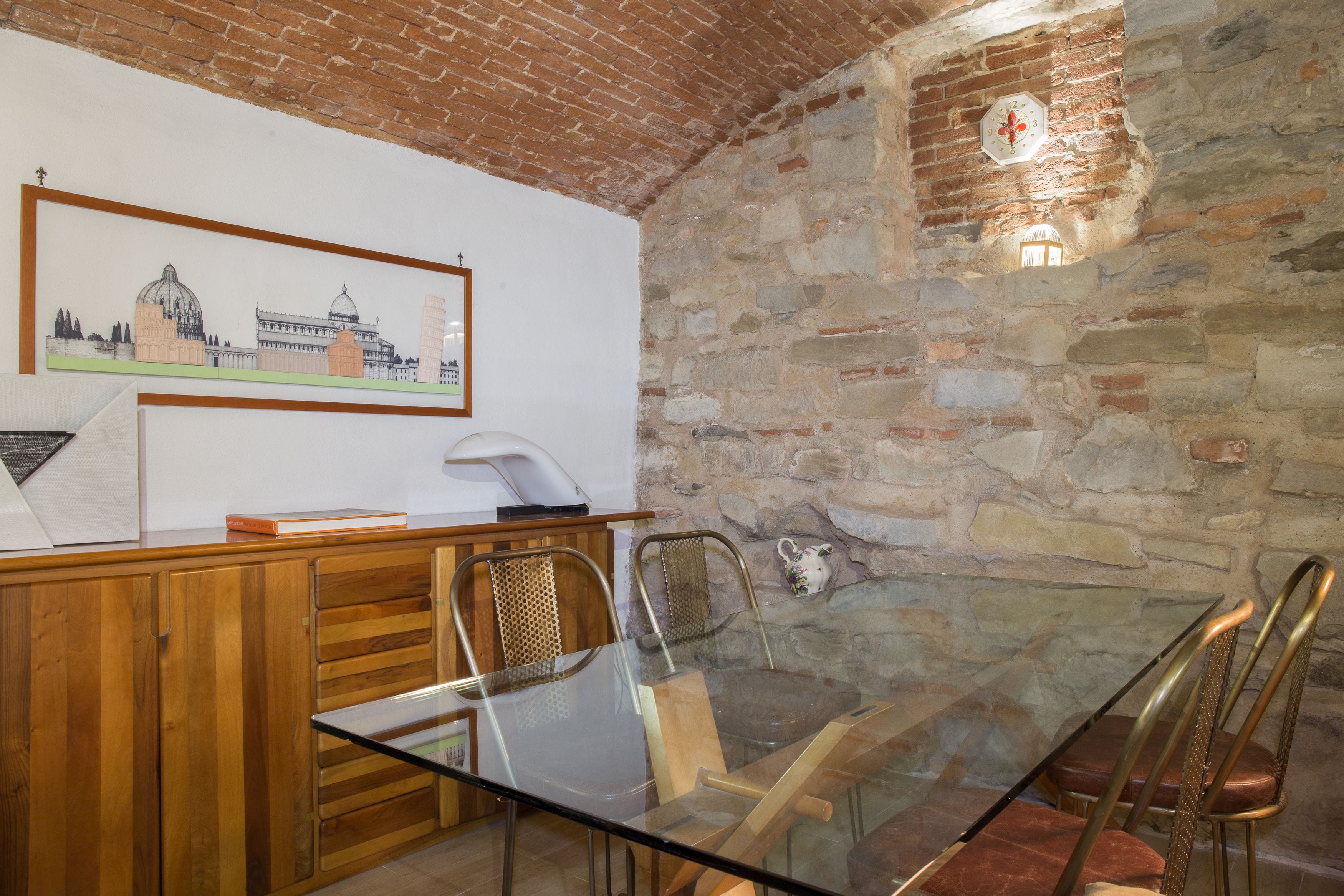 A room with a stone wall, a framed picture of a building, a wooden desk, and a glass table with metal chairs.