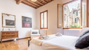 Ein Schlafzimmer mit Holzboden, einem Bett mit weißer Bettwäsche, einem Schminktisch, einem Sessel, einem Heizkörper und einem Fenster mit Blick auf Gebäude.