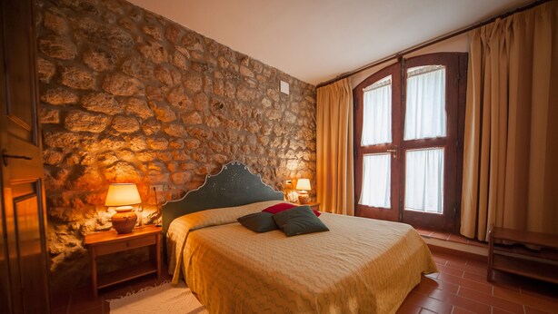 Una camera da letto con parete in pietra, un letto con copriletto decorato, due lampade da comodino e una finestra con tende.