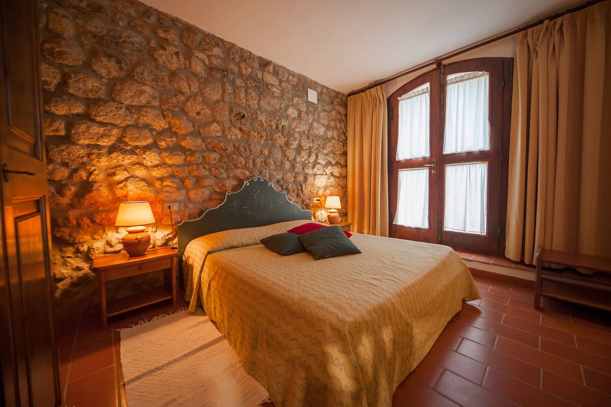 A bedroom with a stone wall, a bed with a patterned bedspread, two bedside lamps, and a window with curtains.