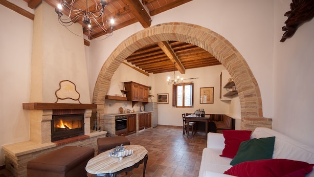 A spacious living area with a fireplace, wooden beams, stone walls, and a white sofa with red and green cushions. - Agriturismo Natura e Salute (San Gimignano)