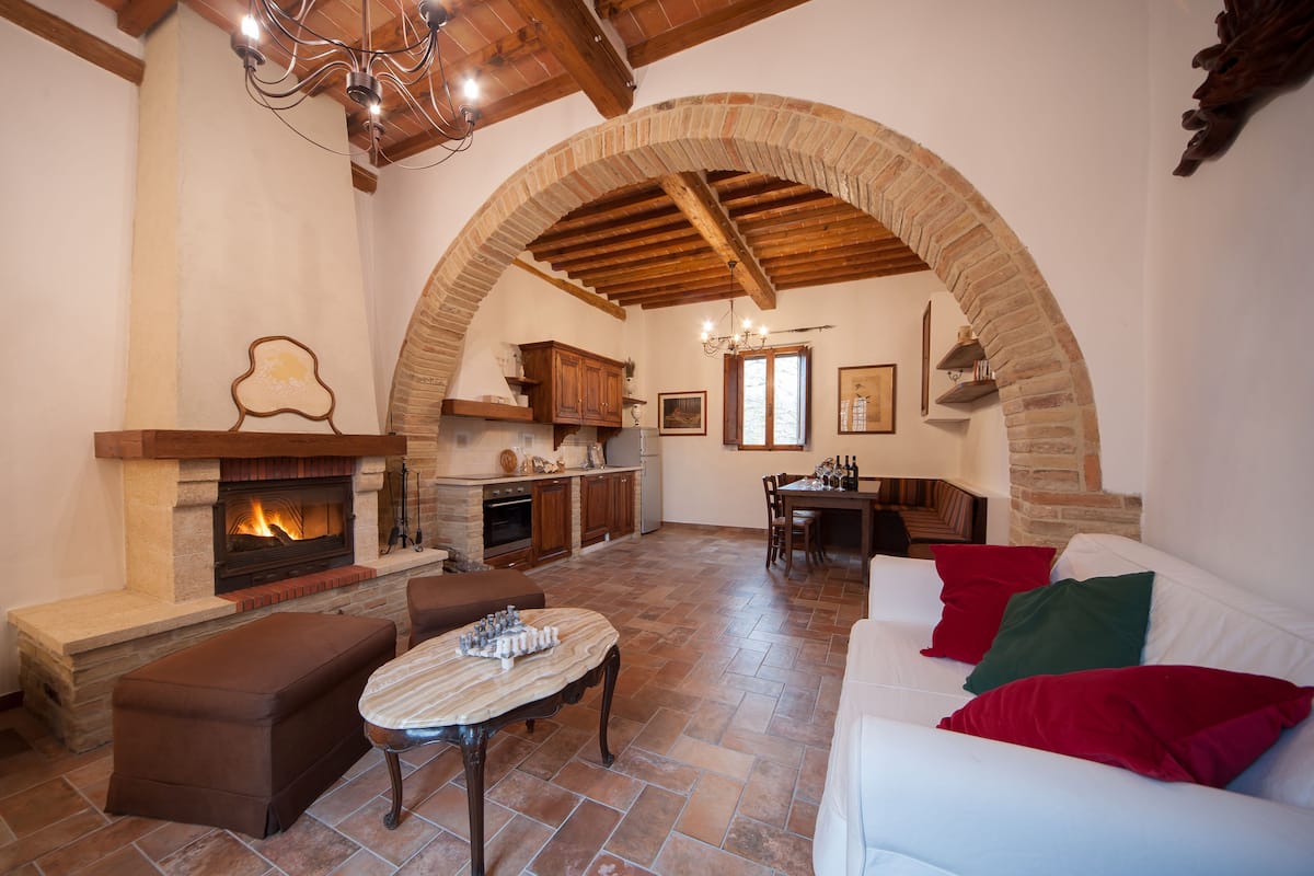 A spacious living area with a fireplace, wooden beams, stone walls, and a white sofa with red and green cushions.